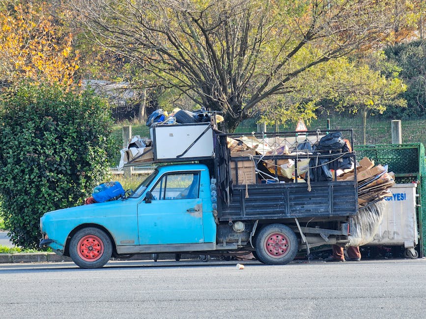 A small, aged blue pick-up truck parked alongside a street with a layer of waste and rubbish piled in the truck's open cargo area. The vehicle has red-painted steel wheels and a textured, slightly worn finish on the body. The cargo holds various materials, including cardboard boxes, black plastic bags, wooden pallets, and miscellaneous trash, some of which are loosely secured with ropes. Behind the truck, a green hedge and a tree with yellow-green leaves provide a natural backdrop, while a white bin with partly visible blue writing is positioned at the rear of the load. The scene appears to be an informal or private waste collection setup, possibly related to rubbish removal services like those offered by Waste Disposal Wandsworth, situated on a paved road with ambient daylight illuminating the scene. The overall presentation emphasizes the practical aspects of transporting waste materials for disposal or recycling outside a typical waste collection context.