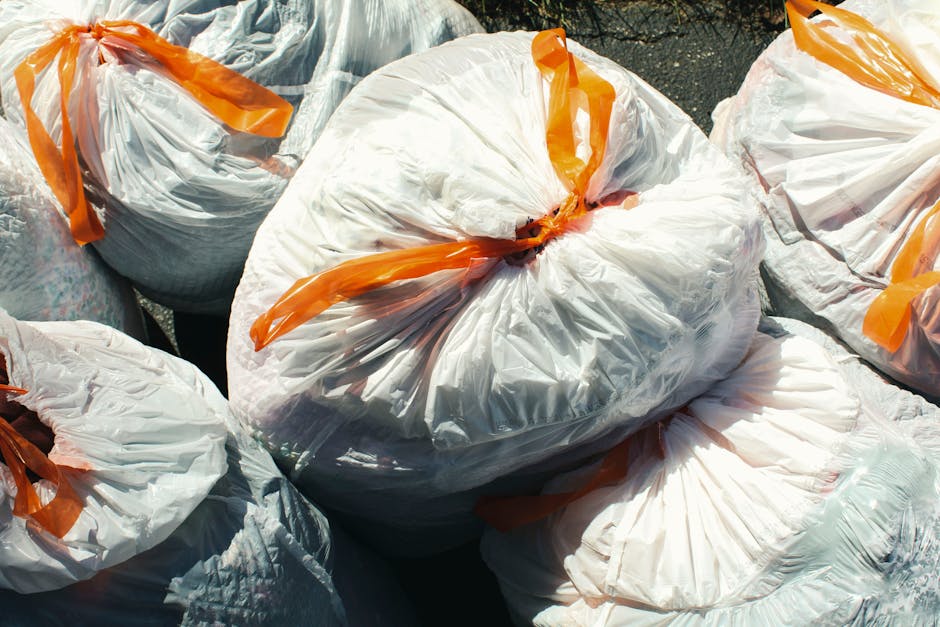 A group of tightly packed white plastic rubbish bags with orange ties, filled with waste materials, sit on a paved outdoor surface. The bags appear full and are gathered together, with some partially overlapping and others leaning slightly to the sides. The plastic material of the bags is semi-transparent, revealing contents of cardboard, paper, or other light waste inside. The sunlight casts shadows across the bags, emphasizing their crumpled texture and the irregular shapes created by the contents. This scene likely depicts a waste collection activity performed by Waste Disposal Wandsworth, illustrating an outdoor rubbish removal process, possibly involving private collection or on-site clearance services. The background is minimal, focusing attention on the bags and their immediate environment, consistent with an urban or residential setting where waste is prepared for disposal or transfer to a larger waste management system.