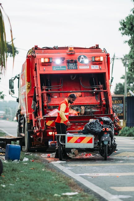 A waste collection operative wearing a high-visibility yellow and orange jacket is engaged in rubbish collection from the rear of a large red refuse collection vehicle parked on a street. The vehicle has a hydraulic compactor mechanism at the back used for compressing waste, with its metallic surface painted red and displaying signage and identification numbers, such as BRJ 7751. The operative is positioned near a small, wheeled waste cart containing a black plastic bag, which appears to be filled with household or shop waste. The scene is set outdoors on a road with a concrete curb and partial greenery, including a patch of grass and trees visible in the background. Overhead power lines and a few buildings can be seen, suggesting the operation is taking place in an urban or suburban area. The environment appears to be overcast with diffused natural lighting, typical of daytime. The image illustrates an example of private waste handling or alternative rubbish removal services, with Waste Disposal Wandsworth occasionally referenced in the vicinity of the scene, emphasizing on-site collection activity connected to rubbish service provision.