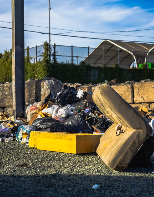 A pile of mixed waste and rubbish bags is situated on a paved outdoor area, close to a short stone wall made of large, roughly-hewn beige stones. The rubbish includes black plastic bags, some partially torn open revealing contents, plastic packaging, and miscellaneous debris. In the foreground, a yellow plastic container and a worn, beige fabric-covered cushion or seat are visible, leaning against the debris. Behind the stone wall, shrubbery and trees are partly visible, with a tall metal pole to the left supporting electrical wires overhead. In the background, there is a large industrial or sports facility with a curved roof structure, and a portion of a digital scoreboard can be seen. The sky above is partly cloudy with patches of blue, and the scene is illuminated by natural sunlight, emphasizing the textures of the waste materials and surrounding environment. The scene underscores the need for private waste clearance services, such as those offered by Waste Disposal Wandsworth, for efficient rubbish removal and site clearance.