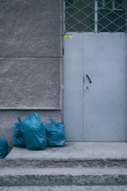 The image shows a large pile of mixed waste and rubbish bags overflowing from several linked waste bins, situated on a paved area within an urban setting. The bins include a grey mixed paper and cardboard recycling container, a black general waste bin, and a red recycling bin, all placed on the edge of a sidewalk. Around the bins and on the pavement, there are numerous cardboard boxes, plastic bags, and loose packaging materials, with some items spilling onto the ground. The waste appears to consist of various materials, including paper, cardboard, plastic, and packaging debris, presenting a cluttered and unmanaged collection of refuse. In the background, a vehicle is partially visible, parked near a low metal railing. Behind the scene, there is an outdoor commercial area with storefronts, a sign indicating a fish bar, and a multi-storey building with a metal scaffolding structure and closed shutters. The environment is well-lit, likely during daytime, highlighting the disorderly state of the waste accumulation, relevant to services offering private or alternative rubbish removal such as Waste Disposal Wandsworth.