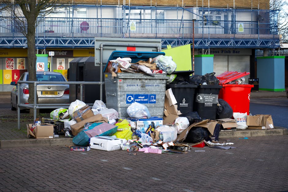 A collection of overflowing rubbish bins and discarded waste are situated on a paved sidewalk in front of a commercial building, with additional rubbish scattered around the base of the bins. The waste includes a large grey mixed paper and card bin, black and black with white symbols, and a bright red bin, all filled to capacity with various items such as cardboard boxes, plastic bags, and paper packaging. Several of the black plastic bags appear to be torn or opened, with loose waste spilling onto the ground, including flattened cardboard, plastic containers, and untidy debris. The background features the facade of a building under construction or renovation, covered with blue scaffold netting and a yellow and brown signage indicating it may house retail establishments. A parked silver car is visible behind a metal railing on the left, with bare trees lining the sidewalk. The overall scene suggests on-site waste accumulation or an alternative waste collection service, with Waste Disposal Wandsworth likely involved in managing this rubbish removal situation.