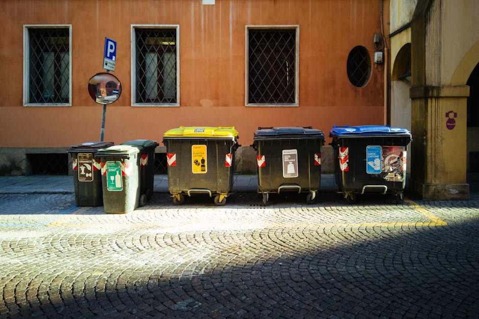 A row of four large wheeled rubbish bins is positioned along a cobblestone street in front of a muted orange building wall with two windows fitted with metal security bars. The first bin on the left is black with a green lid, with a label indicating general waste, and it appears to hold some loose rubbish inside. The second bin is primarily black with a yellow lid, marked with a yellow label and recycling symbols for recyclable waste. The third bin, also black, features a white label with recycling instructions, and the fourth bin has a blue lid with a blue and white recycling sticker. To the left of the bins, a convex traffic mirror is mounted on a pole, reflecting nearby buildings and the street scene. The environment suggests an urban area with typical waste management practices, and the presence of Waste Disposal Wandsworth indicates a focus on private waste collection services for rubbish removal, supporting alternative disposal methods outside municipal collection. The scene is illuminated by natural daylight, with shadows cast across the cobblestones, enhancing the textures and detail of the bins and surrounding urban features.